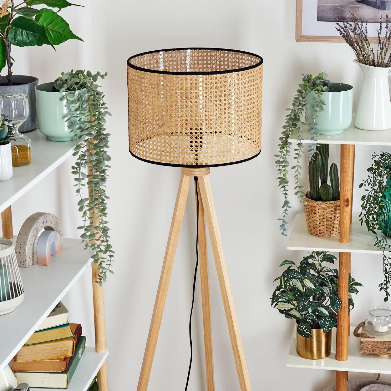 Eine Holz-Dreibein-Lampe mit gewebtem Lampenschirm steht vor weißen Regalen mit Pflanzen und Dekorationsartikeln.