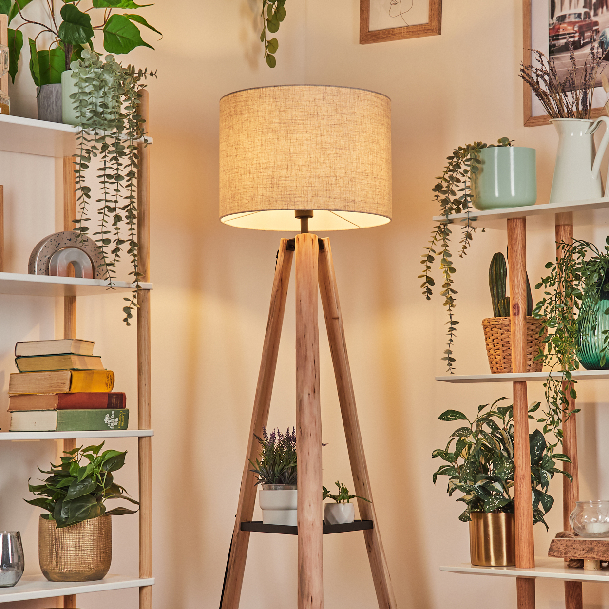 Eine Holz-Dreibein-Lampe steht in einem Raum mit Regalen voller Pflanzen und Bücher. Die Lampe hat einen beigen Lampenschirm.