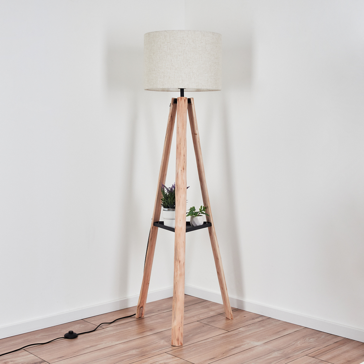 Eine hölzerne Stehlampe mit einem beigefarbenen Lampenschirm. Sie hat ein schwarzes Regal mit Topfpflanzen. Der Hintergrund ist eine weiße Wand und Holzboden.