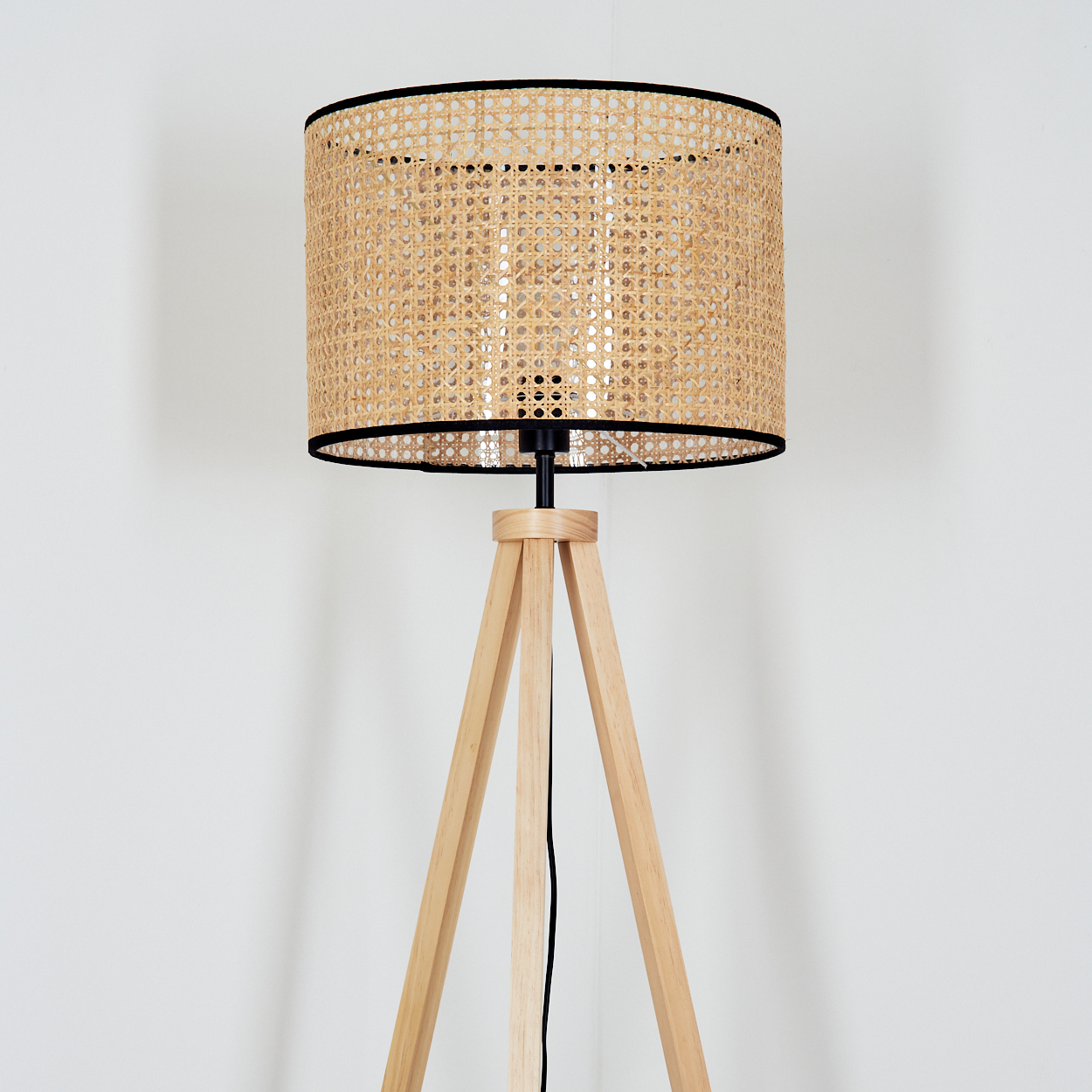 Eine Stehlampe mit gewebtem Lampenschirm und Holzstativbeinen steht vor einer weißen Wand. Ein schwarzes Kabel ist sichtbar.