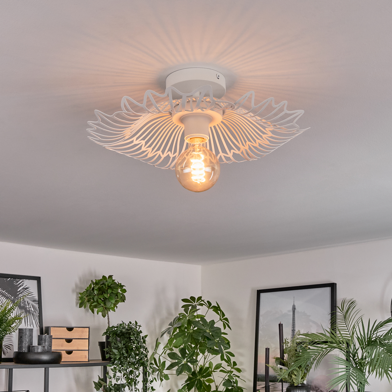 Eine weiße Deckenlampe mit dekorativem Design, die einen Raum mit Pflanzen und gerahmten Bildern beleuchtet.