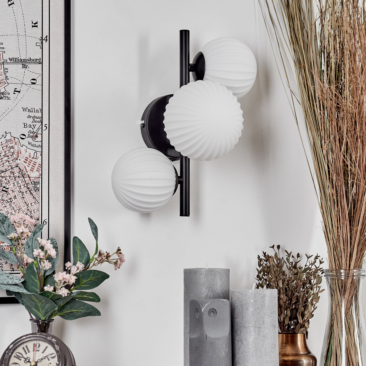 Eine moderne Wandlampe mit drei weißen Kugelleuchten. Daneben Kerzen, getrocknete Pflanzen in einer Vase und eine Karte.