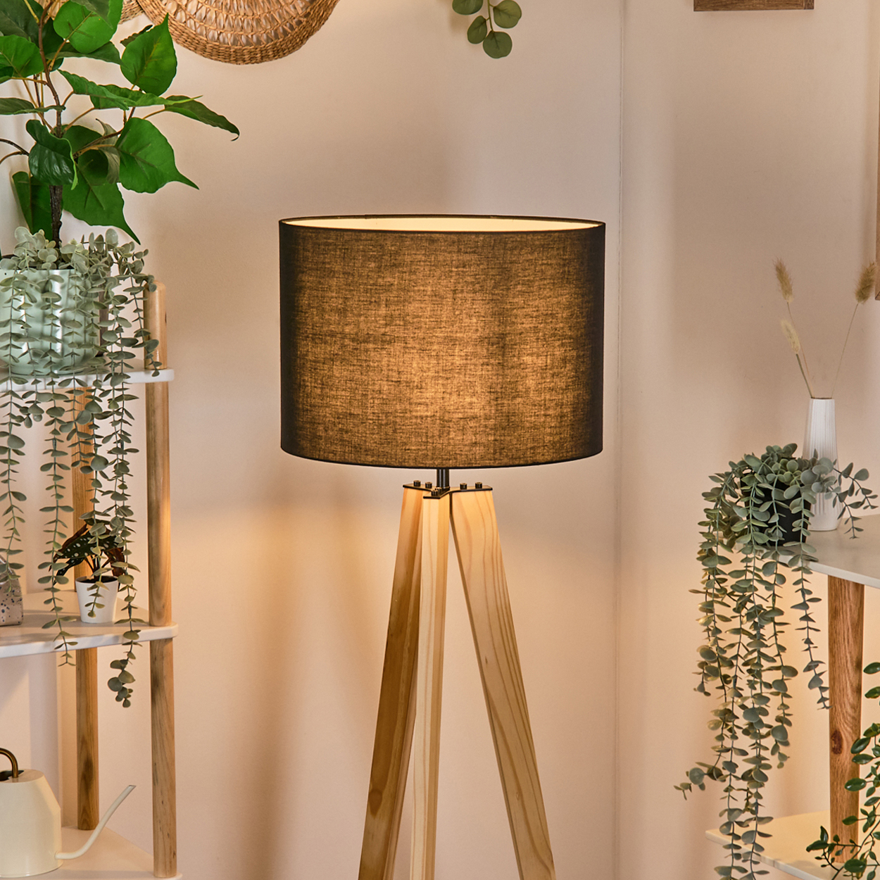 Eine hölzerne Stehlampe mit schwarzem Lampenschirm. Pflanzen und Regale befinden sich im Hintergrund. Eine weiße Wand vervollständigt die Szene.