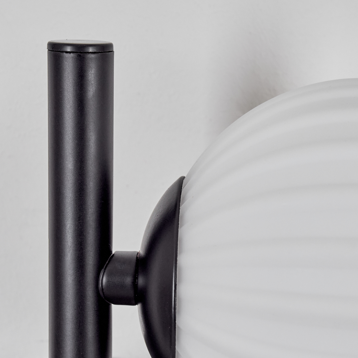 Nahaufnahme einer Lampe mit schwarzem Metallrahmen und weißem, geripptem Glasschirm. Der Hintergrund ist eine weiße Wand.