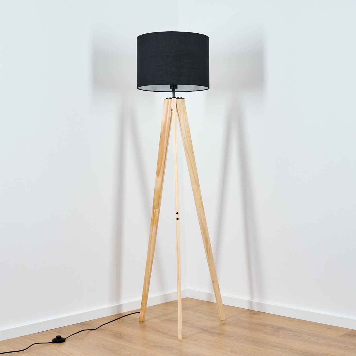 Eine Stehlampe mit Holzstativ und schwarzem Lampenschirm steht in einer Ecke. Der Raum hat weiße Wände und einen Holzboden.