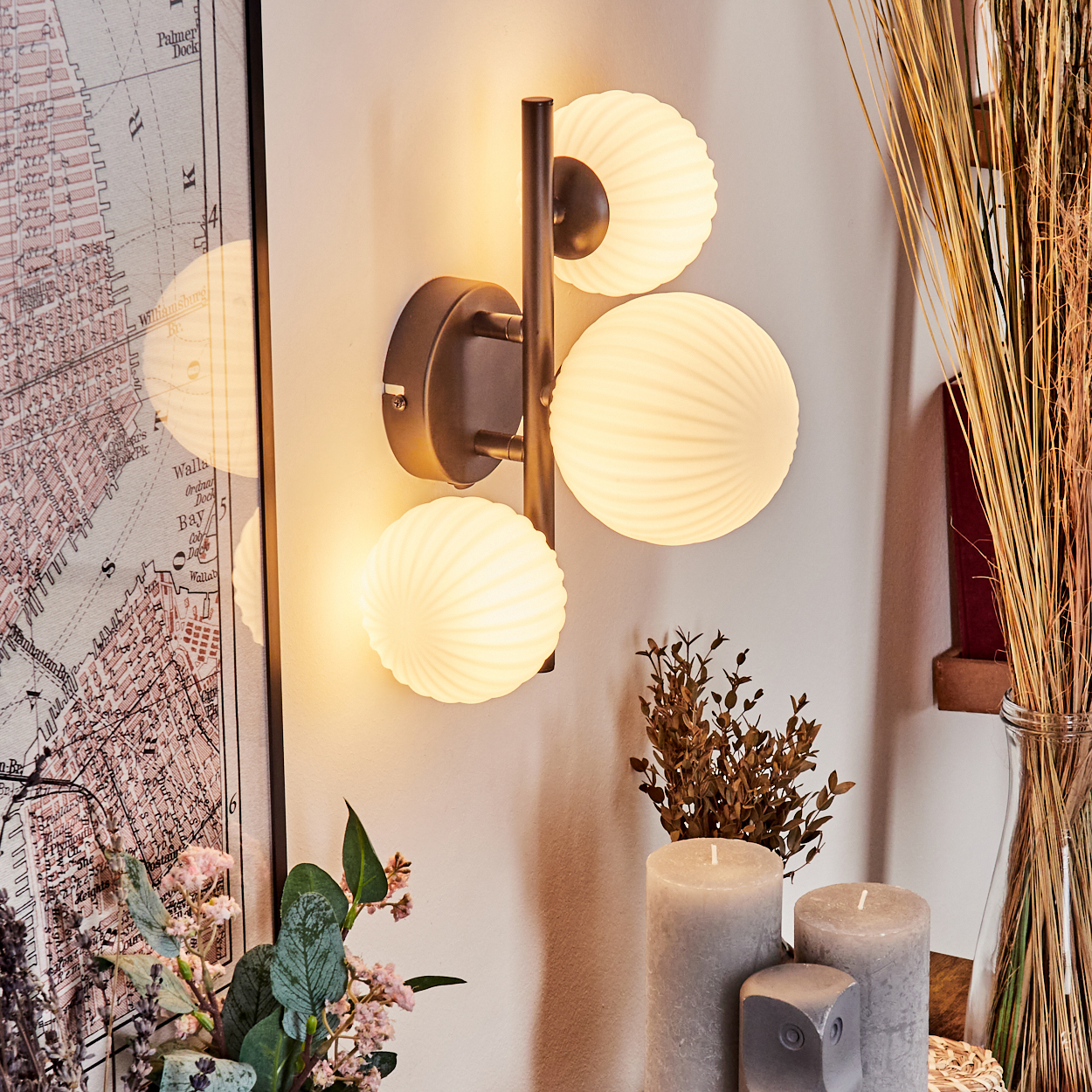 Eine Wandlampe mit drei Kugelleuchten. Dahinter sind eine Karte und Pflanzen zu sehen. Davor graue Kerzen und getrocknete Pflanzen.