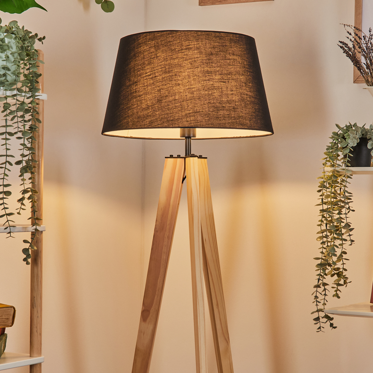 Eine hölzerne Stativ-Stehlampe mit schwarzem Lampenschirm steht in einem Raum. Im Hintergrund sind grüne Pflanzen und weiße Regale zu sehen.