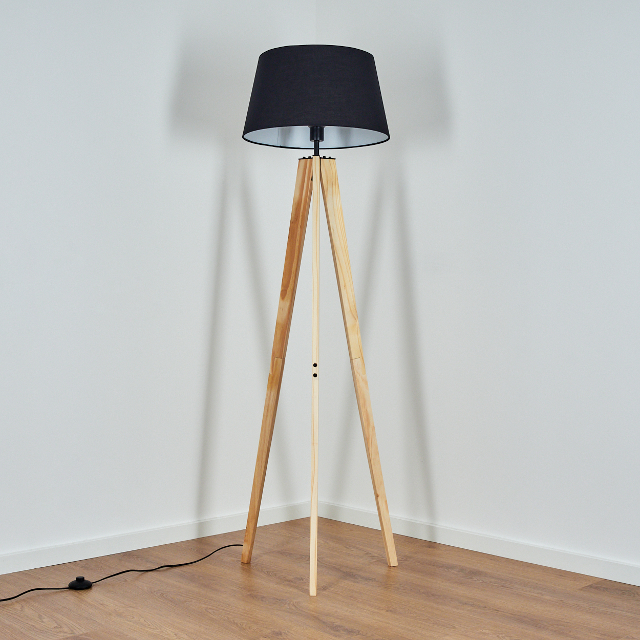 Eine hölzerne Stativ-Stehlampe mit schwarzem Lampenschirm steht in einer Ecke. Der Boden ist aus Holz, die Wände sind weiß.