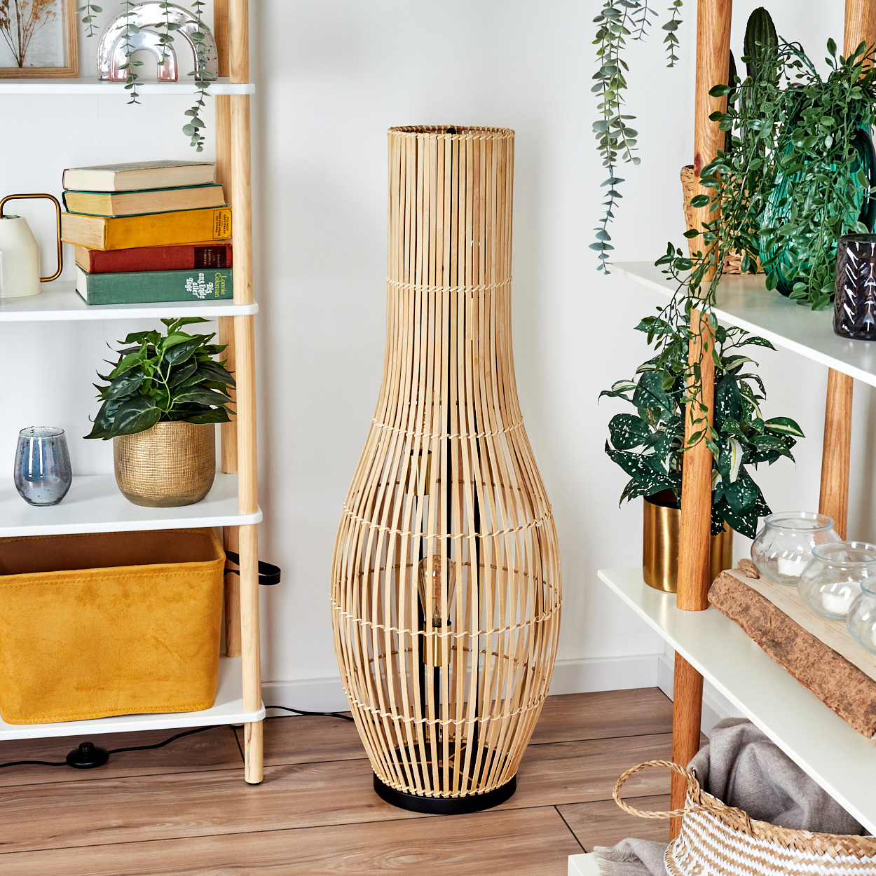 Eine hohe, gewebte Bambuslampe steht in einem Raum mit Regalen voller Pflanzen und Bücher. Der Boden ist aus Holz.