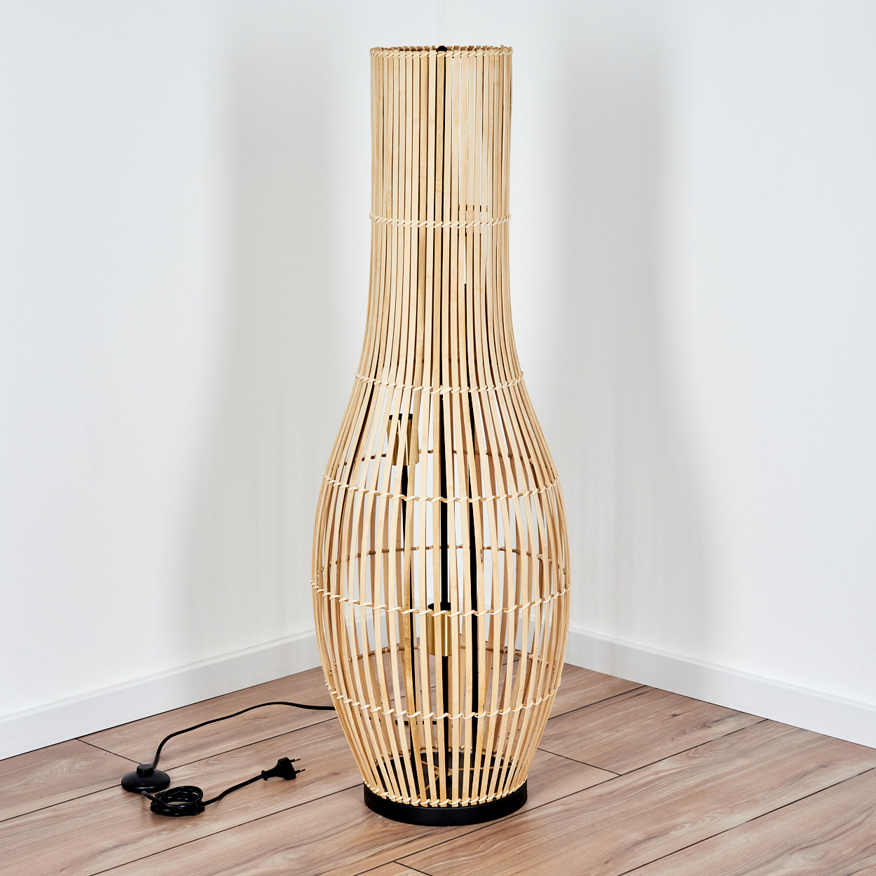 Eine hohe, beige Stehlampe aus Korbgeflecht steht in einer Ecke. Die Lampe hat einen schwarzen Sockel und ein Kabel. Der Hintergrund ist weiß und hat einen Holzboden.