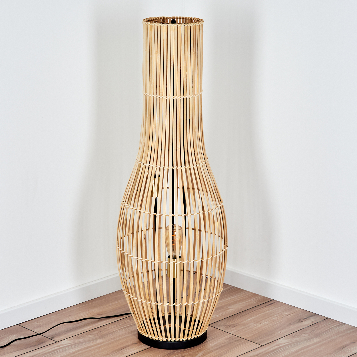 Eine hohe, gewebte Stehlampe aus Bambus steht in einer Ecke. Die Lampe ist beige, hat einen schwarzen Sockel und eine sichtbare Glühbirne.