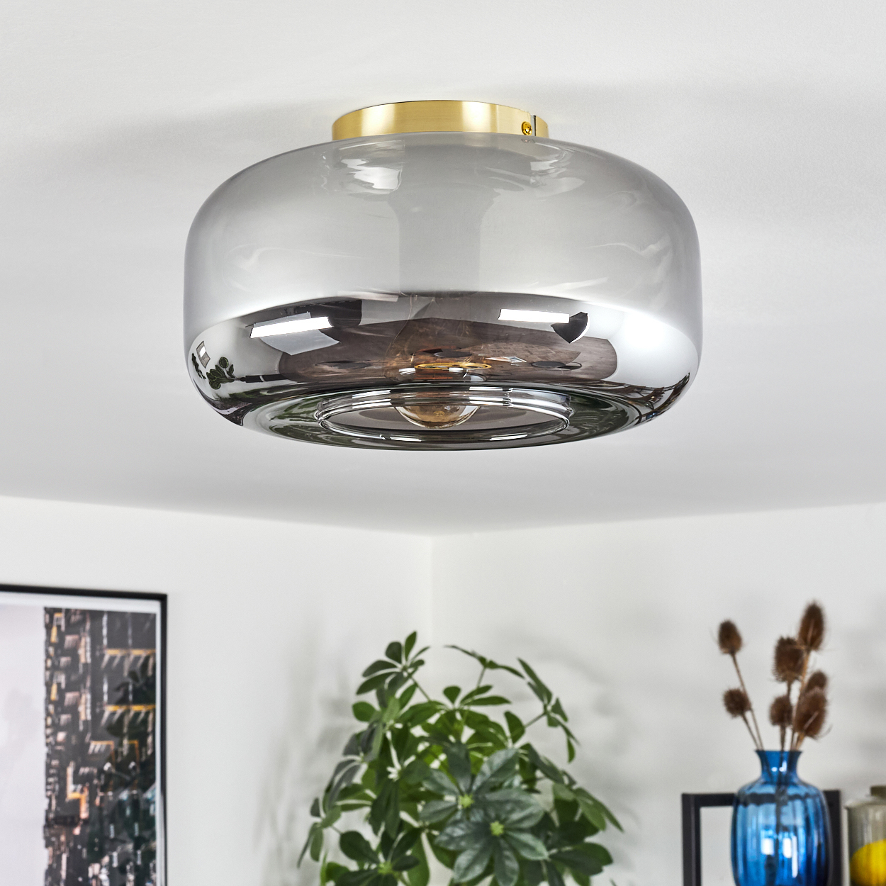 Eine moderne Deckenleuchte mit goldenem Oberteil und grauem, reflektierendem Glasschirm. Pflanzen und eine blaue Vase im Hintergrund.