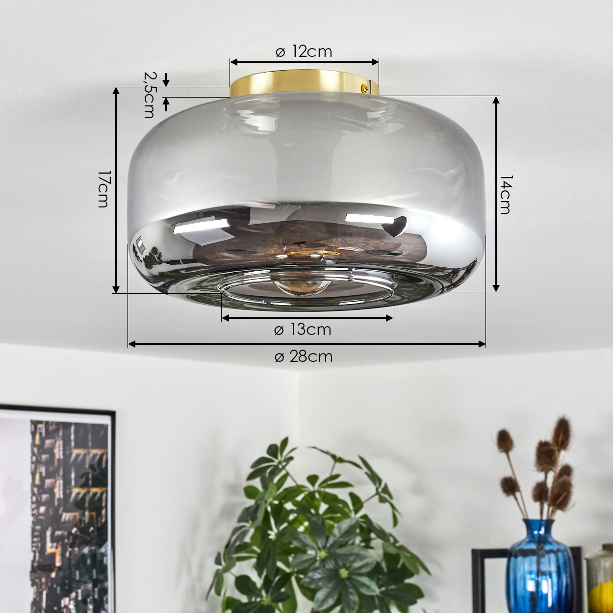 Eine moderne Deckenlampe mit goldenem Sockel und rauchfarbenem Glasschirm. Maße sind markiert. Pflanzen und eine blaue Vase im Hintergrund.