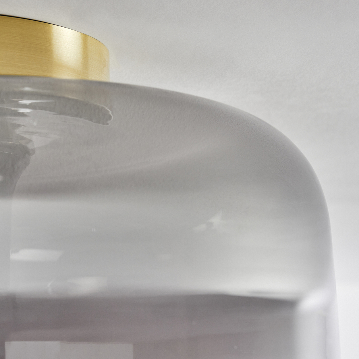 Nahaufnahme einer Leuchte. Sie hat einen goldfarbenen Deckel und einen grauen, abgerundeten Glasschirm. Der Hintergrund ist weiß.