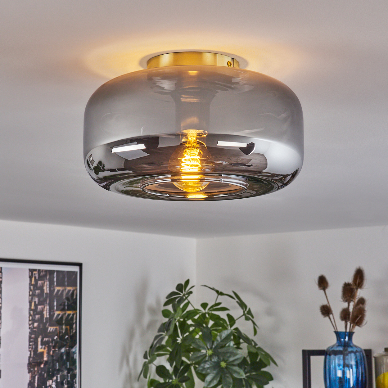 Deckenleuchte mit grauem Glasschirm und goldenem Sockel. Eine leuchtende Glühbirne ist innen. Pflanzen und ein Bilderrahmen sind im Hintergrund.