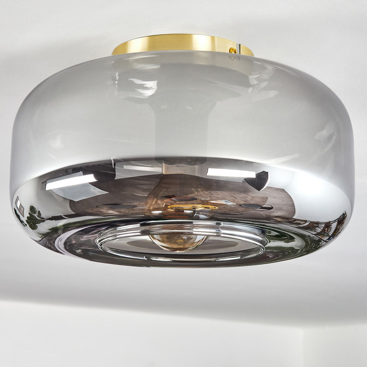 Eine moderne Deckenlampe mit grauem Glasschirm und goldfarbenem Metallsockel. Die Lampe spiegelt die Umgebung wider.