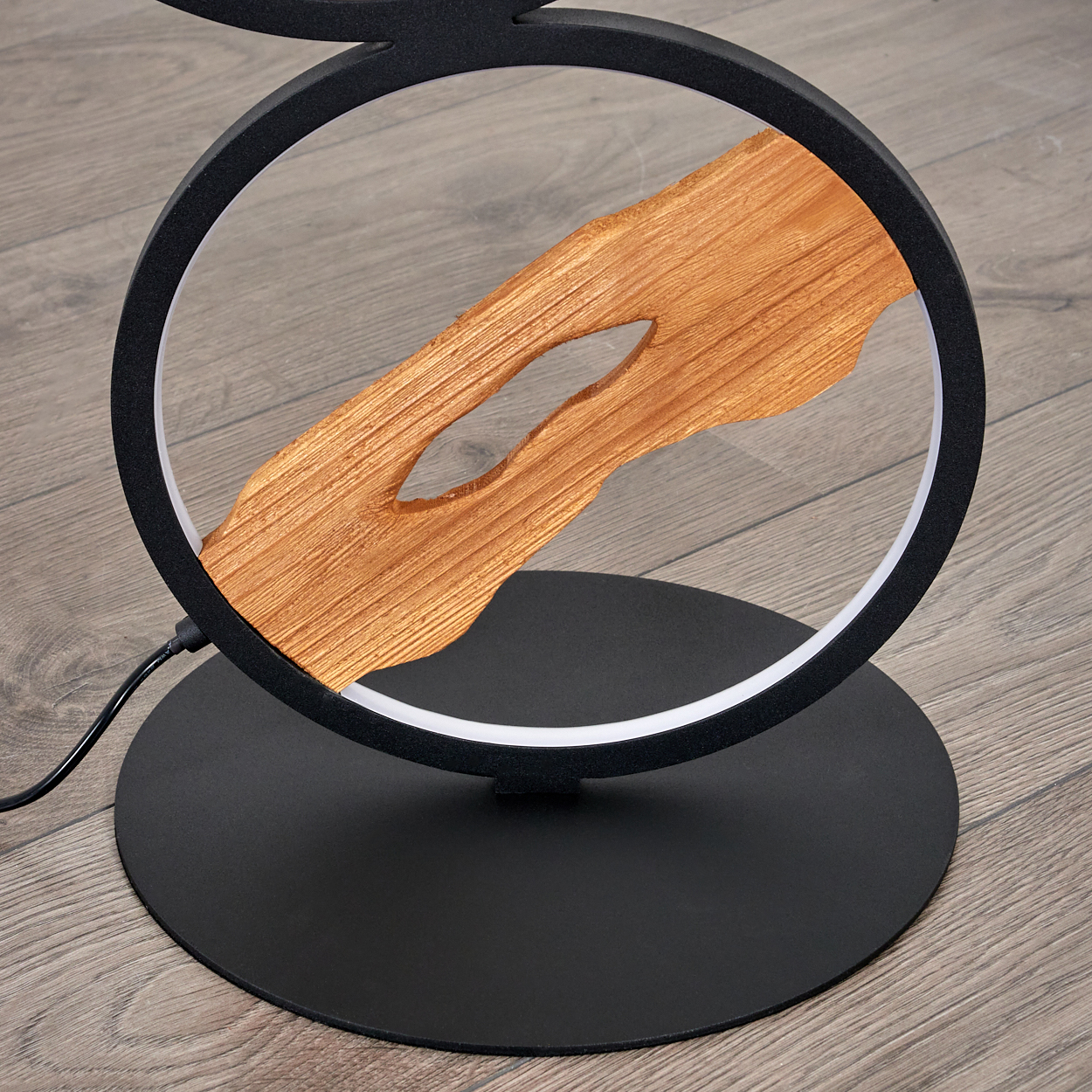 Eine moderne Lampe mit einem Holzmittelstück in einem schwarzen kreisförmigen Rahmen auf einem schwarzen Sockel. Der Hintergrund ist ein Holzboden.