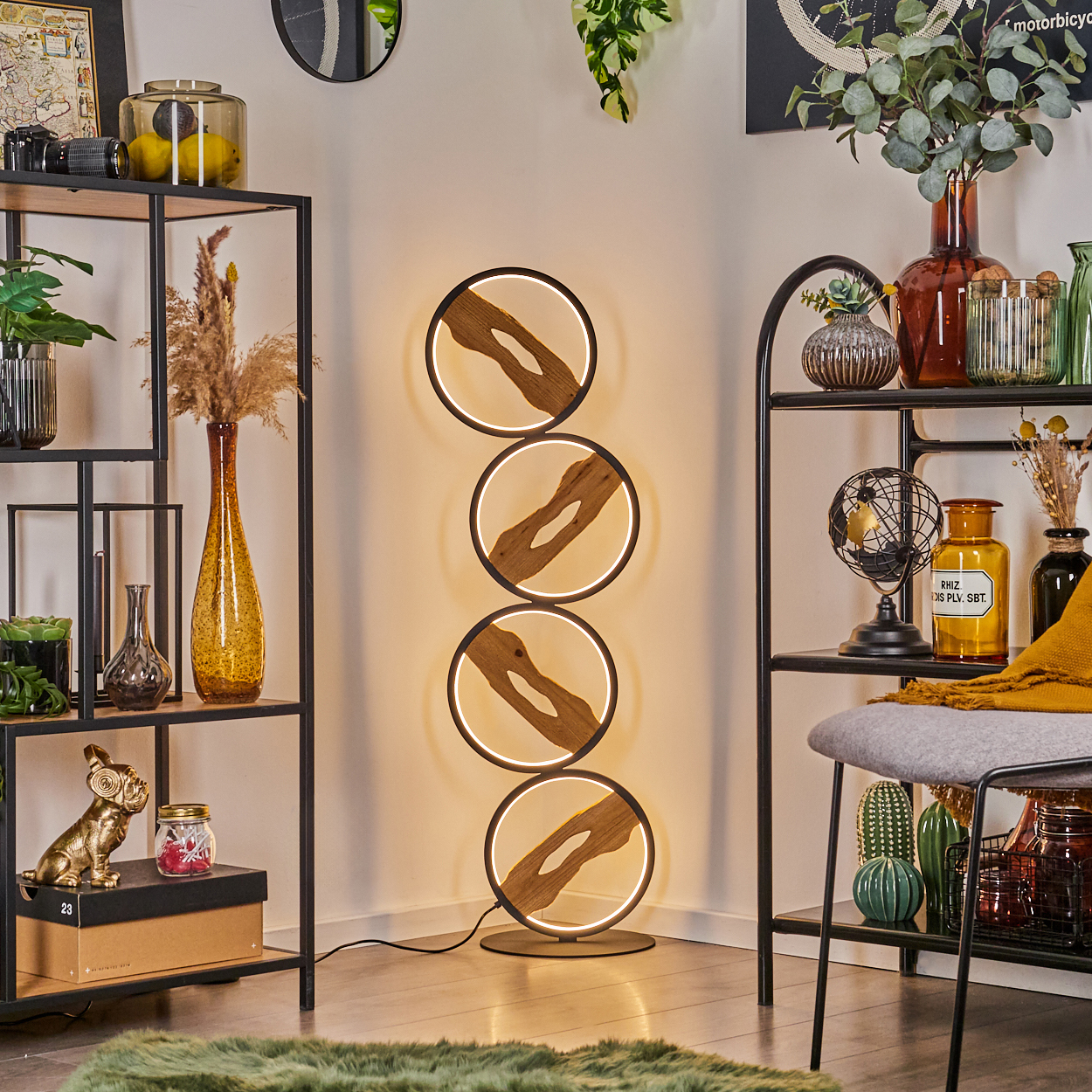 Ein modernes Wohnzimmer mit einer Stehlampe aus vier kreisförmigen Lichtern. Regale zeigen Dekorationen, ein grüner Teppich liegt auf dem Boden.