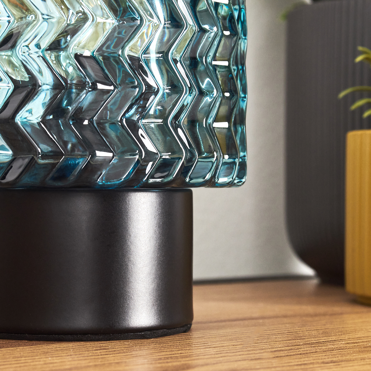 Nahaufnahme einer tealfarbenen Glasvase mit schwarzem Sockel. Der Hintergrund umfasst eine dunkelgraue und eine gelbe Vase.