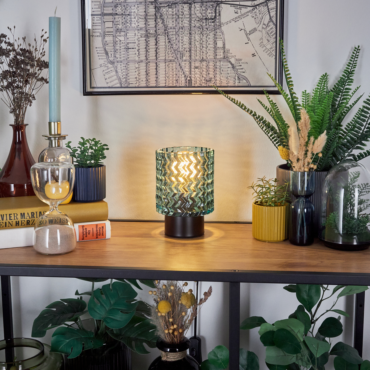 Ein Tisch mit Dekoration: eine Lampe, Pflanzen, eine Sanduhr, eine Kerze, Bücher und eine Karte. Die Lampe ist grün, die Pflanzen sind vielfältig und die Karte ist schwarz-weiß.