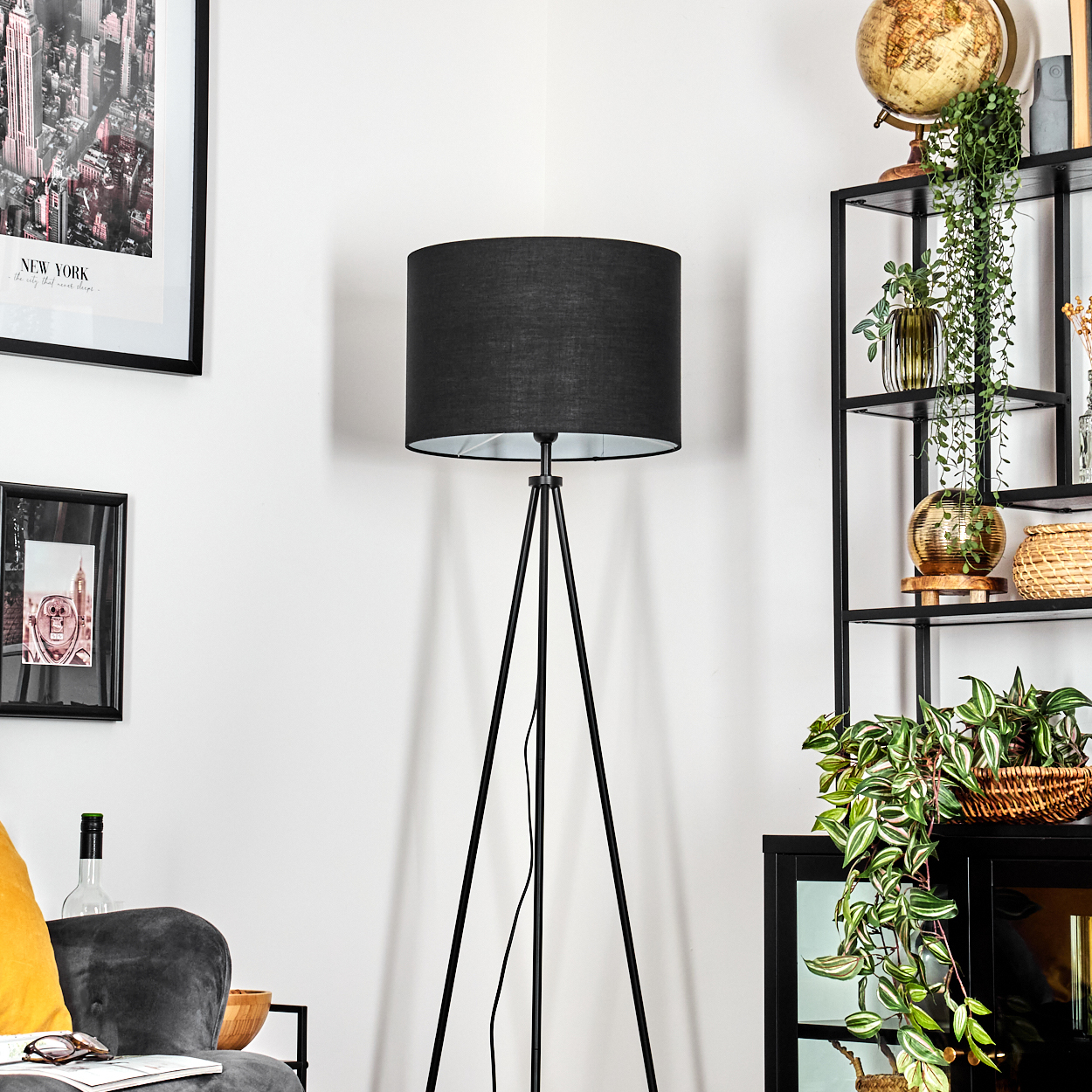Eine schwarze Stehlampe steht in einer Ecke. Rechts ein schwarzes Regal mit Pflanzen und einem Globus. Bilder hängen an der Wand.
