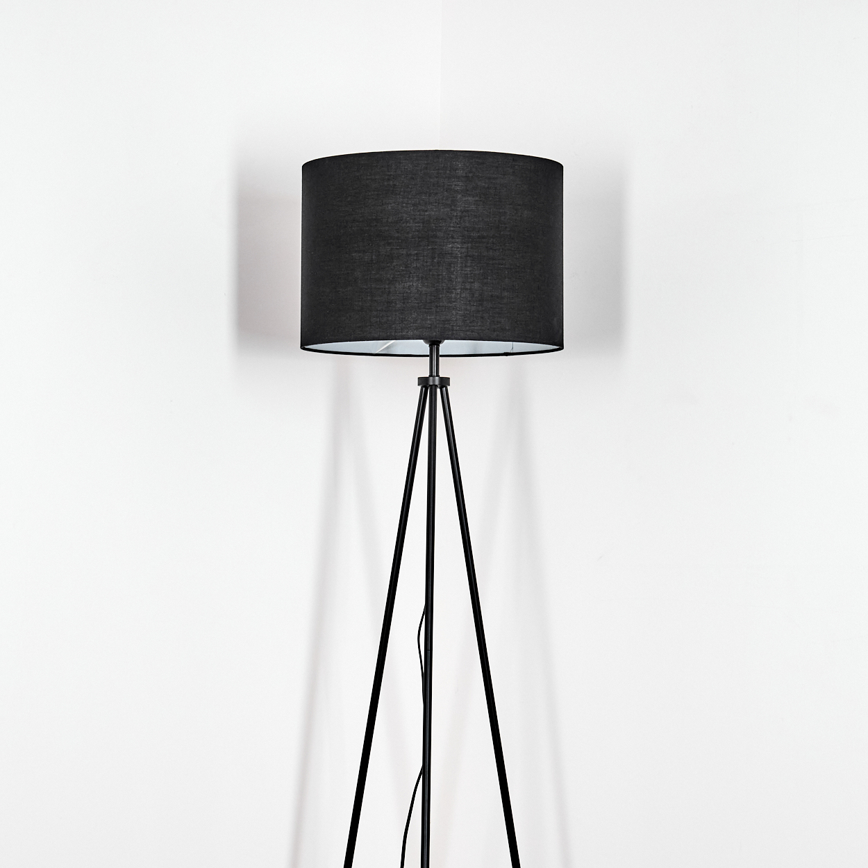 Eine schwarze Stehlampe mit zylindrischem Lampenschirm steht vor einer weißen Wand. Die Beine der Lampe sind im Dreibein-Stil.