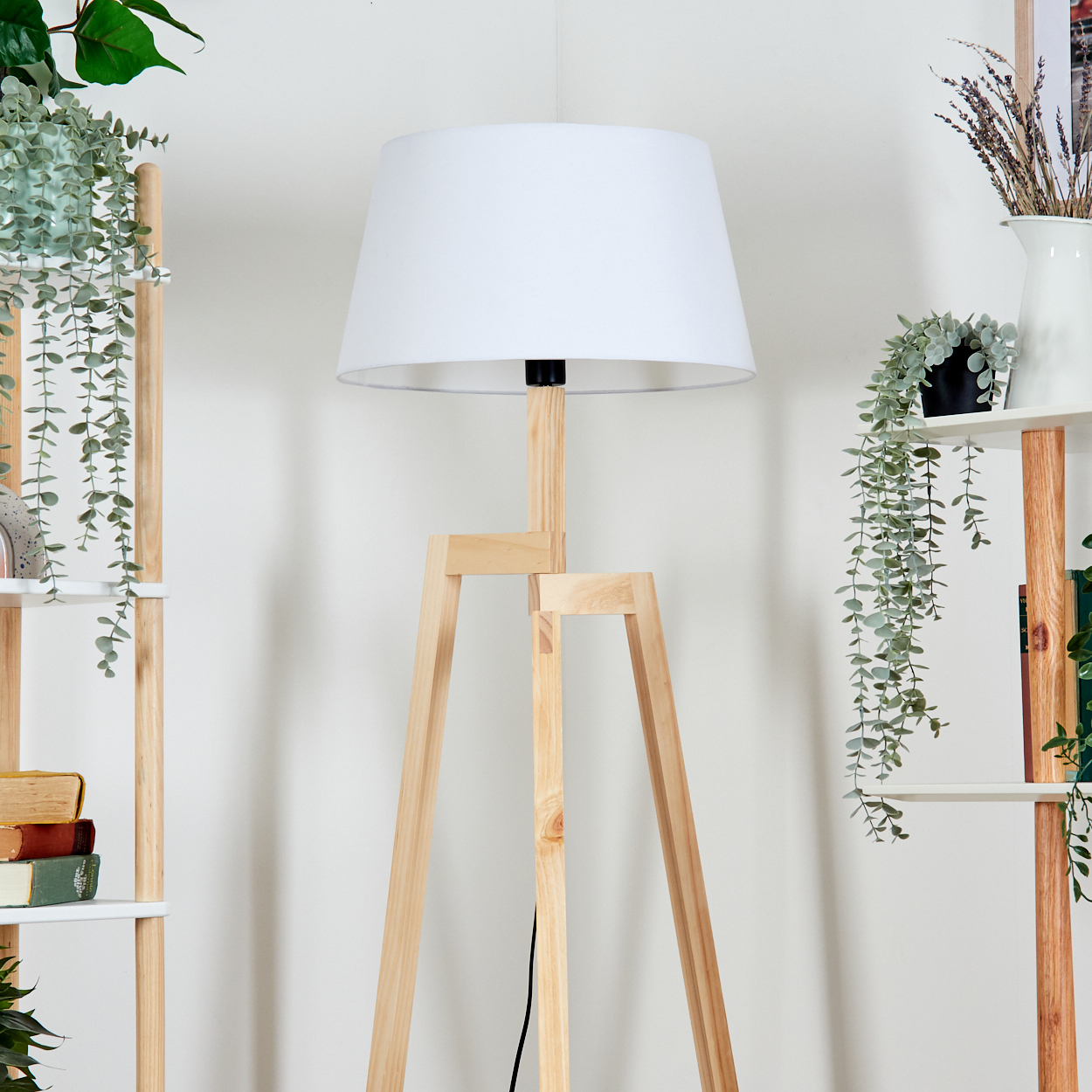 Eine hölzerne Stativlampe mit weißem Lampenschirm steht vor einer weißen Wand. Regale mit Pflanzen und Büchern sind an den Seiten.