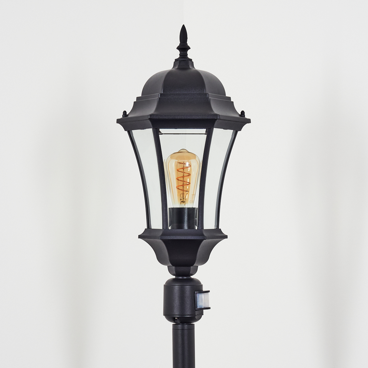Schwarze Außenlampe mit einer Glühbirne im Vintage-Stil. Die Lampe befindet sich auf einer schwarzen Stange mit Bewegungssensor. Weißer Hintergrund.