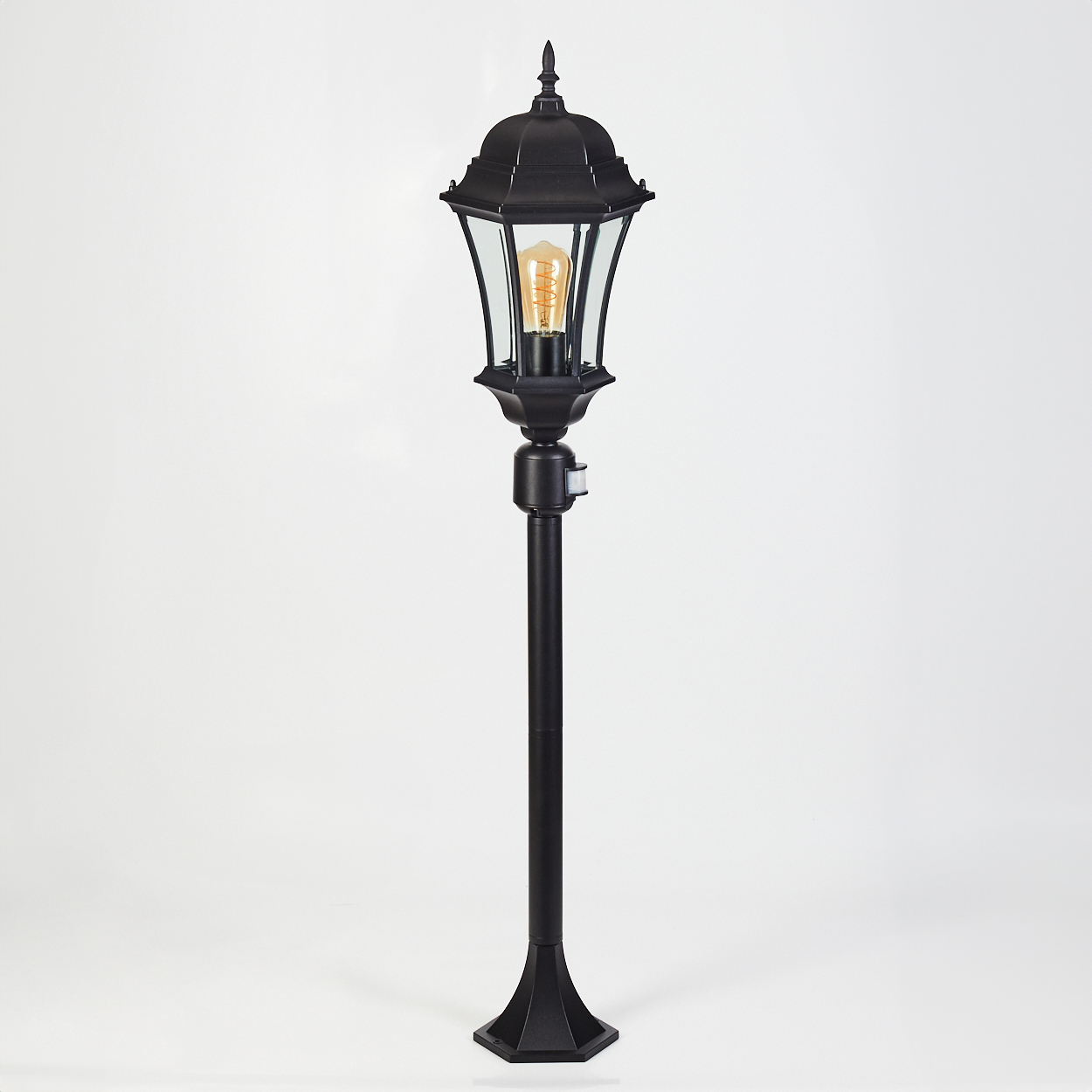 Schwarze Außenlampe auf weißem Hintergrund. Sie hat ein Glasgehäuse, eine warme Glühbirne und einen Bewegungsmelder.