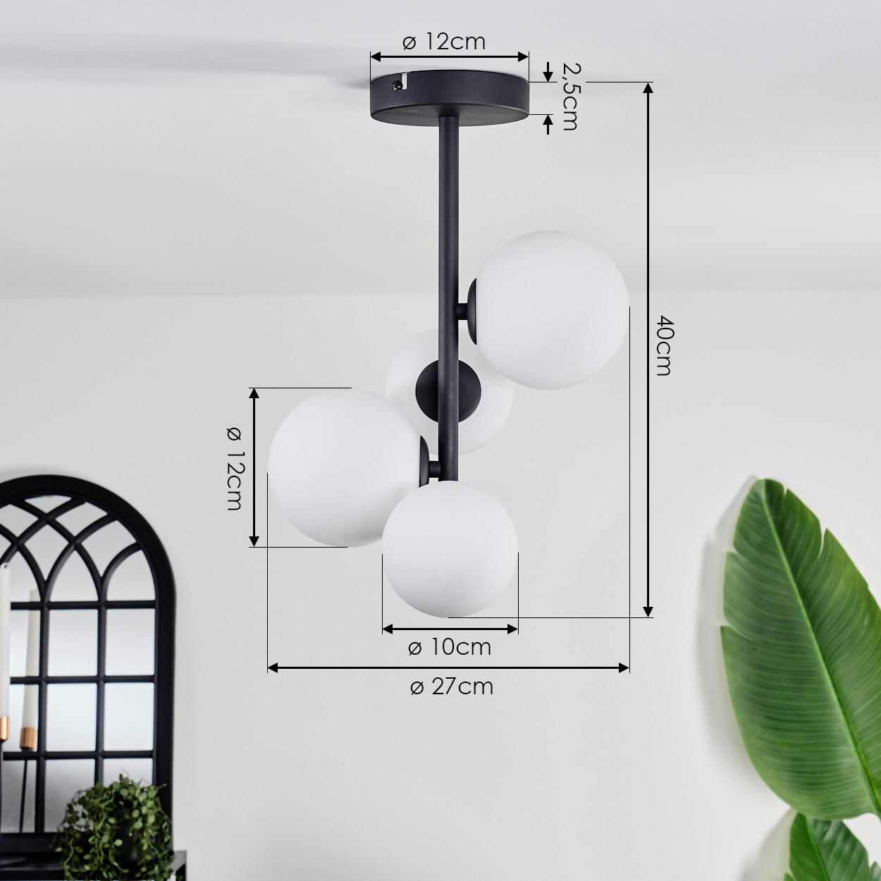 Eine schwarz-weiße Deckenlampe mit vier kugelförmigen Glühbirnen. Die Lampe befindet sich vor einer weißen Wand, in der Nähe eines Spiegels und einer Pflanze.
