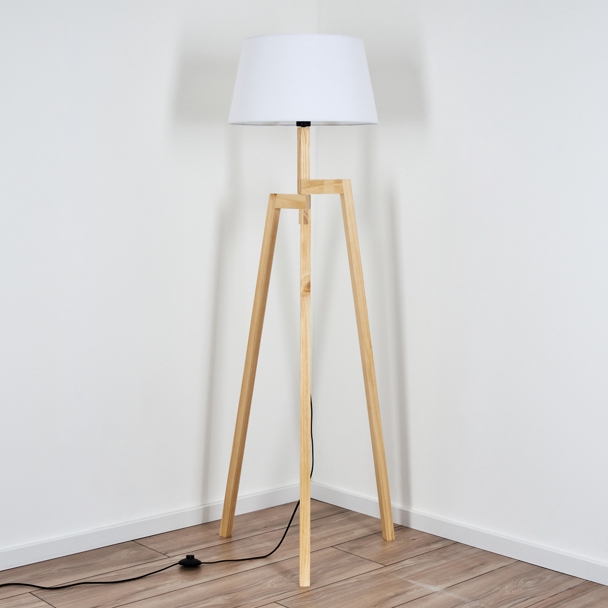 Eine hölzerne Stativ-Stehlampe mit weißem Lampenschirm steht in einer Ecke auf einem Holzboden vor einer weißen Wand.