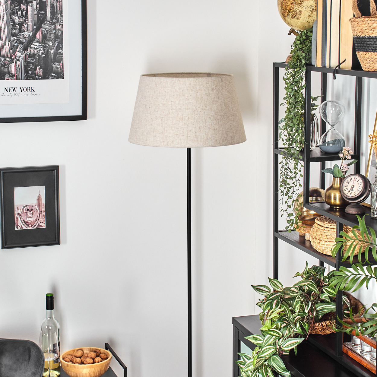 Eine Stehlampe mit beigem Schirm steht vor einer weißen Wand. Ein Regal mit Deko, ein gerahmtes Bild und eine Schüssel mit Nüssen sind sichtbar.