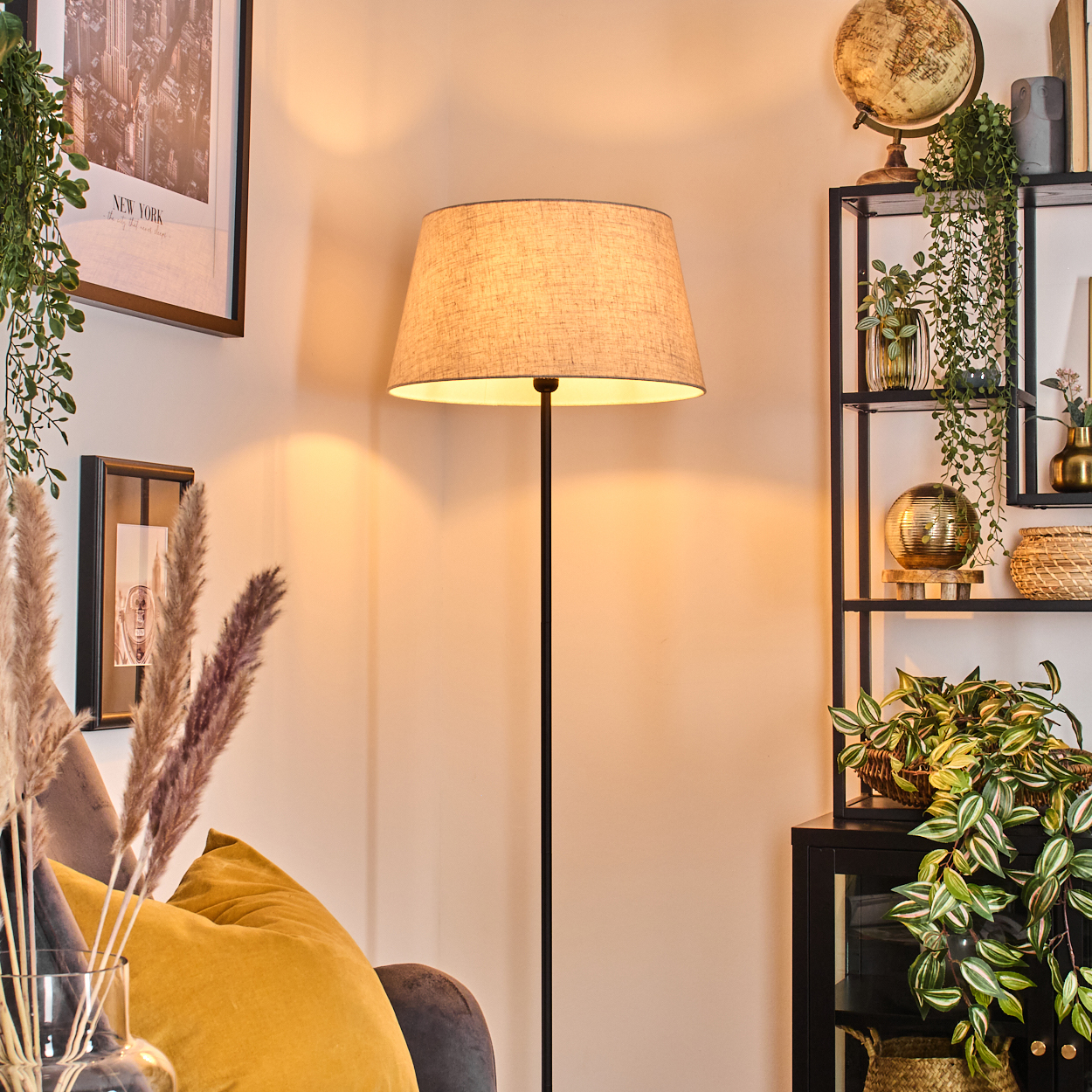 Eine beleuchtete Lampe in einem Raum mit Dekoration, darunter ein Globus, Pflanzen und ein gerahmtes Bild. Links befindet sich ein gelbes Kissen.