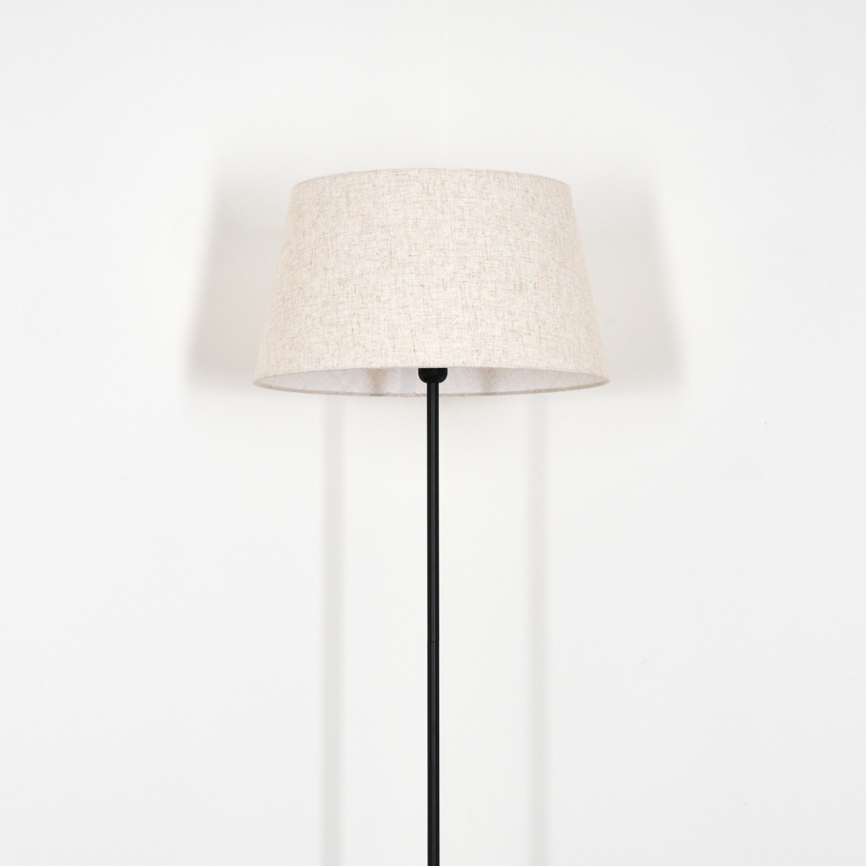 Eine Stehlampe mit beigem Lampenschirm steht vor einer weißen Wand. Die Lampe hat einen schwarzen Pfosten. Der Schatten ist an der Wand sichtbar.