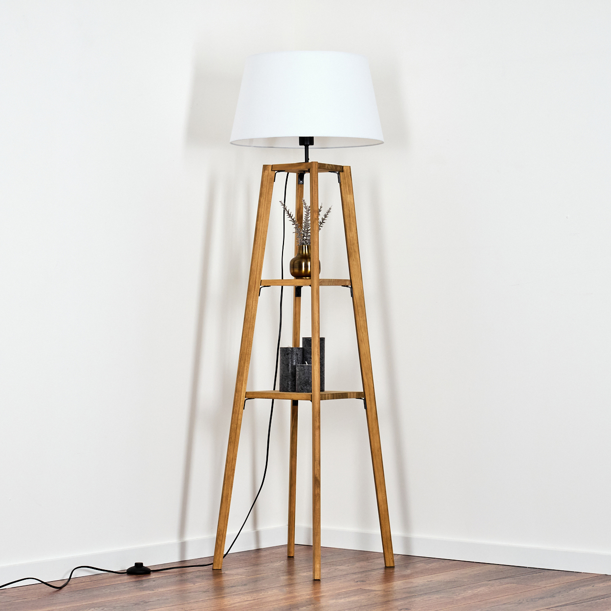 Eine Stehlampe aus Holz mit weißem Lampenschirm. Sie hat Regale mit einer goldenen Vase mit Pflanzen und schwarzen Kerzen. Die Wände sind weiß.