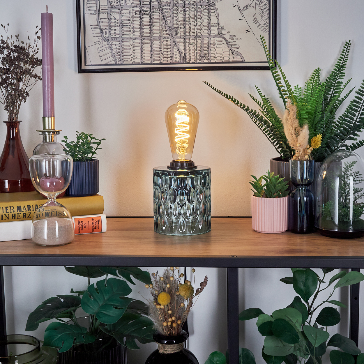 Ein Holzregal mit Lampe, Pflanzen und Dekoration. Eine Karte hängt an der weißen Wand. Eine Kerze, eine Sanduhr und Bücher sind ebenfalls vorhanden.