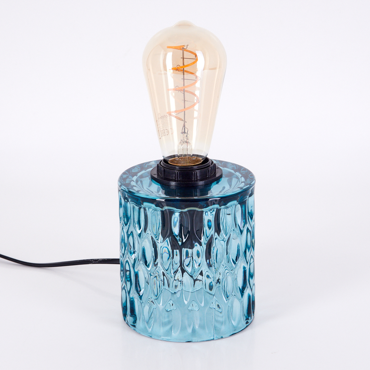 Eine dekorative Tischlampe mit blauem Glassockel und Edison-Glühbirne. Der Sockel hat ein strukturiertes Muster. Ein schwarzes Kabel erstreckt sich.