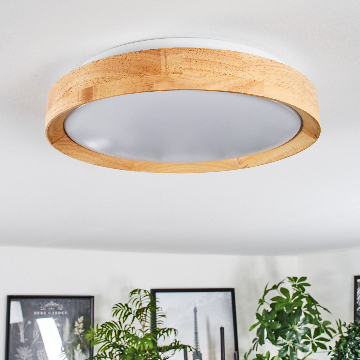 Eine Deckenleuchte aus Holz und Weiß ist an einer weißen Decke montiert. Im Hintergrund befinden sich grüne Pflanzen und gerahmte Bilder.