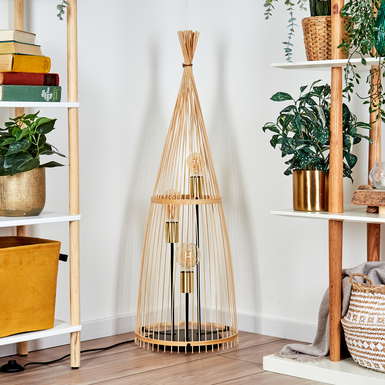 Eine Bambuslampe mit drei Glühbirnen steht vor Regalen mit Pflanzen und Büchern. Der Raum hat eine weiße Wand und einen Holzboden.