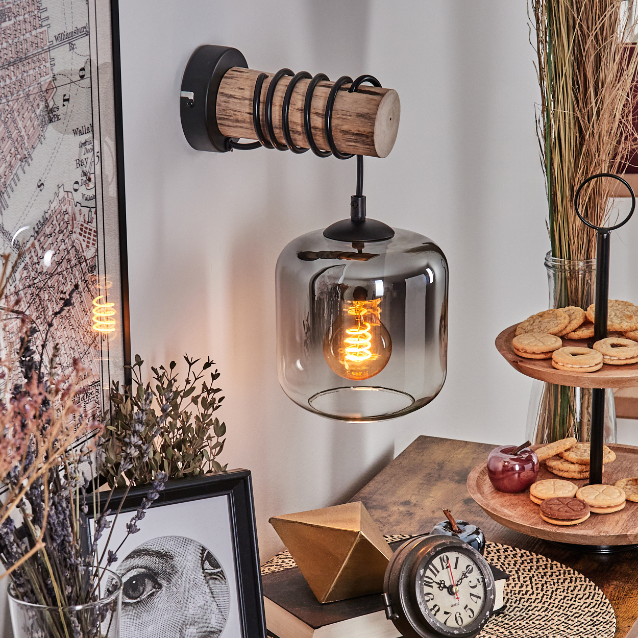Eine Wandlampe mit Holz- und Metalldesign. Es gibt einen Glasschirm mit einer sichtbaren Glühbirne. Kekse, Bücher und ein Augenkunstwerk befinden sich in der Nähe.