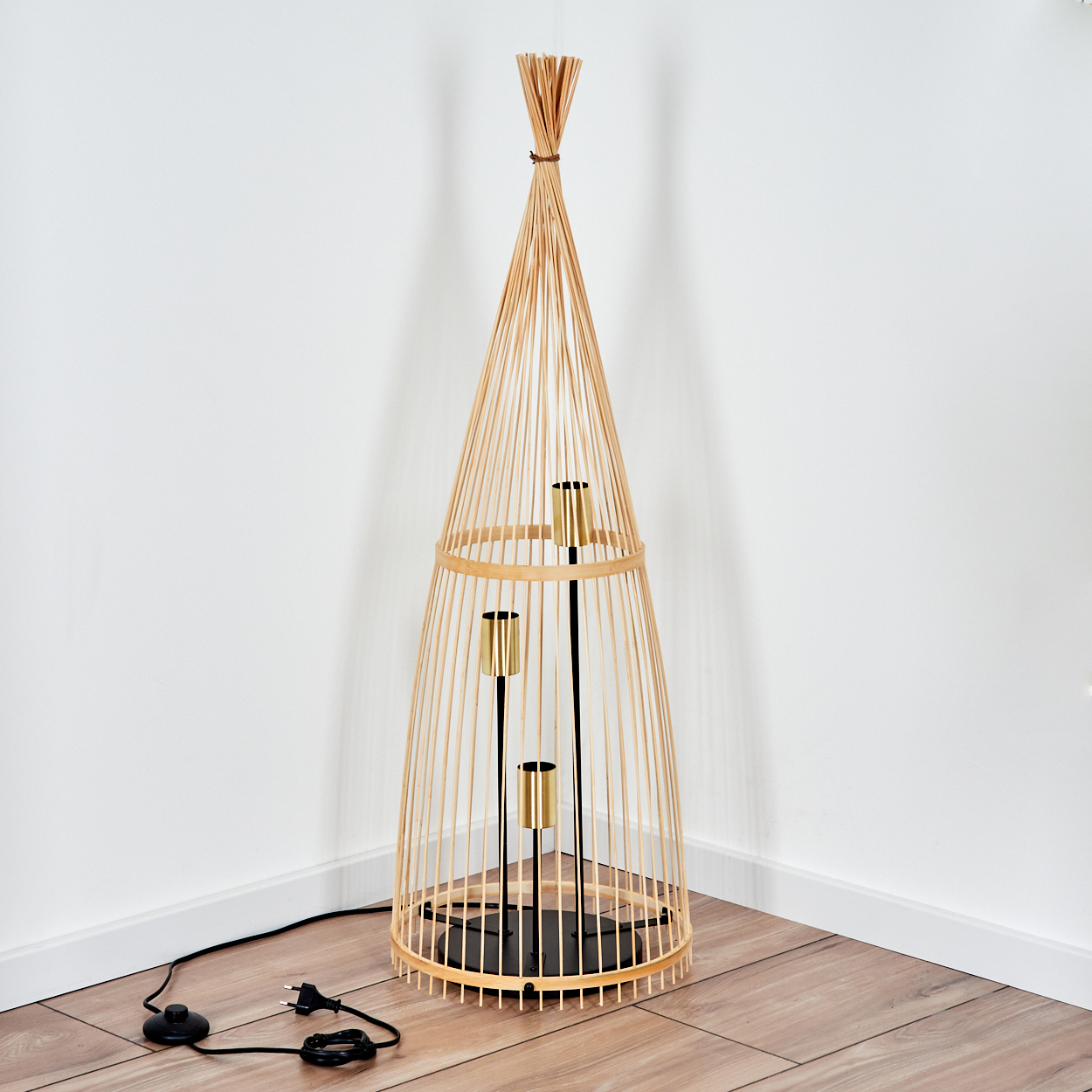 Eine Bambus-Stehlampe mit drei goldenen Lichthaltern steht in einer Zimmerecke. Netzkabel und Stecker liegen auf dem Holzboden.