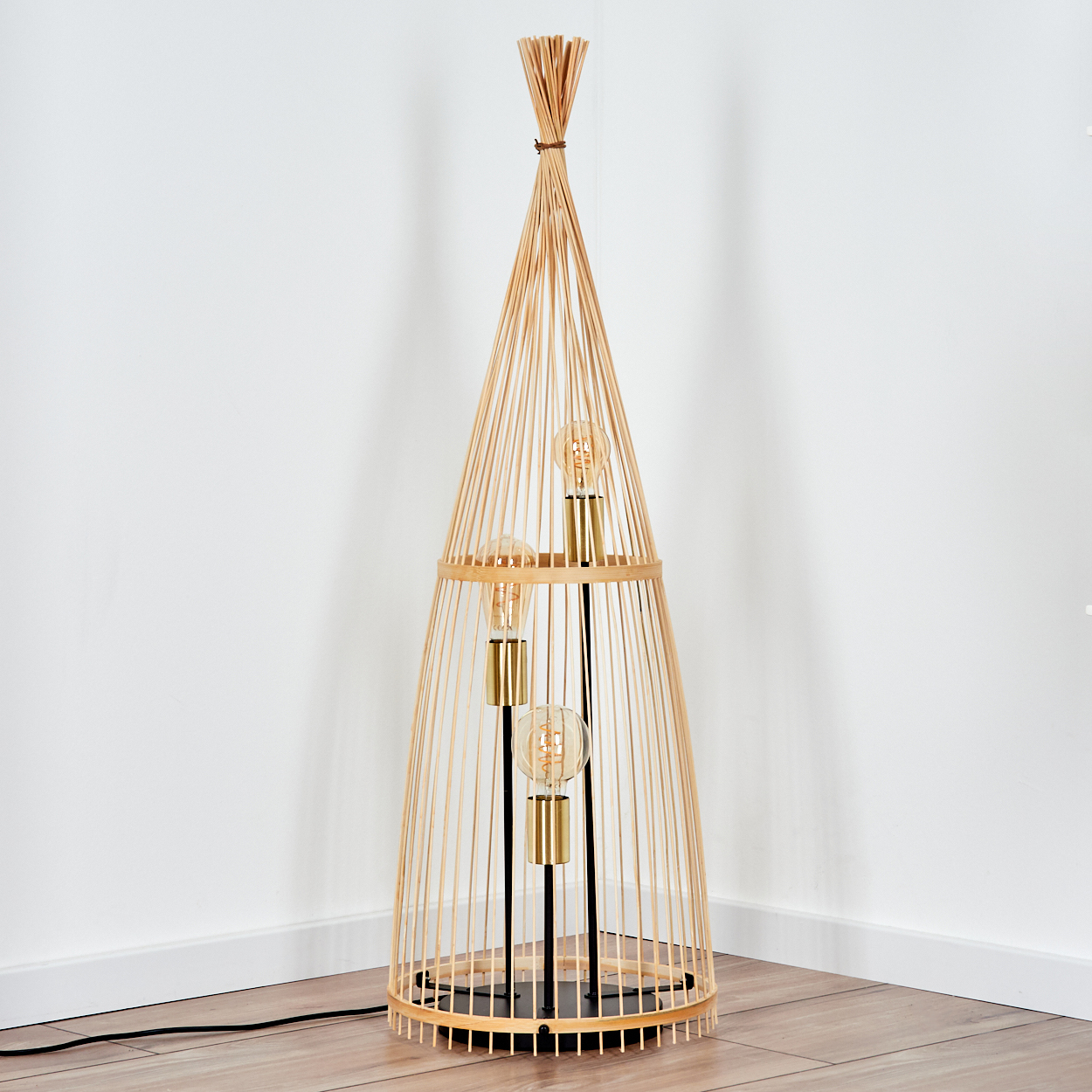 Eine hohe Stehlampe aus Bambus mit vier Glühbirnen, die in einer Ecke steht. Der Hintergrund ist eine weiße Wand und ein Holzboden.