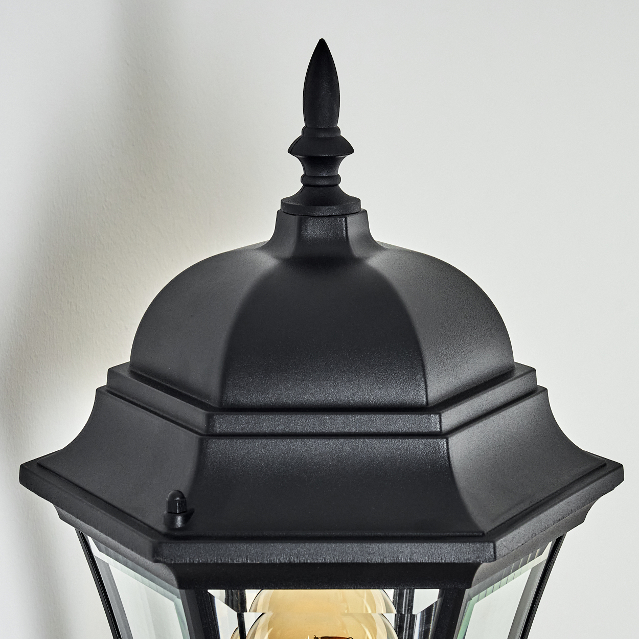 Nahaufnahme einer schwarzen Außenlampe. Sie hat eine spitze Spitze, ein gewölbtes Dach und Glasscheiben. Der Hintergrund ist eine weiße Wand.