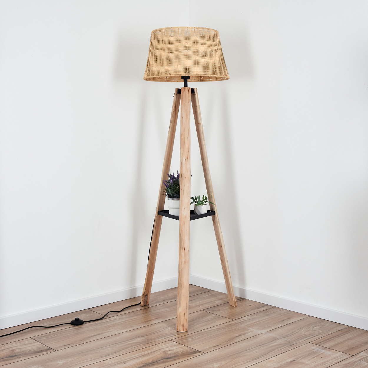 Eine Stehlampe mit gewebtem Lampenschirm und Holzstativ. Zwei kleine Topfpflanzen stehen auf einem Regal. Der Hintergrund ist eine weiße Wand.