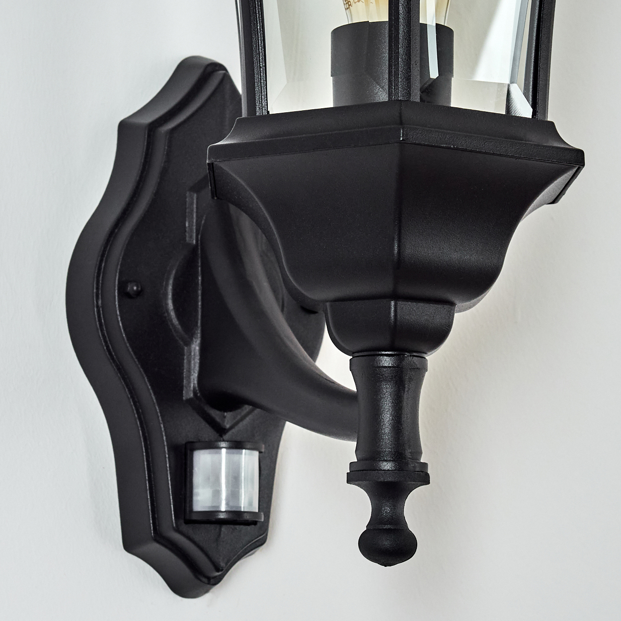 Eine schwarze Außenwandleuchte mit Glasgehäuse. Sie hat einen Bewegungssensor. Die Leuchte ist an einer weißen Wand montiert.