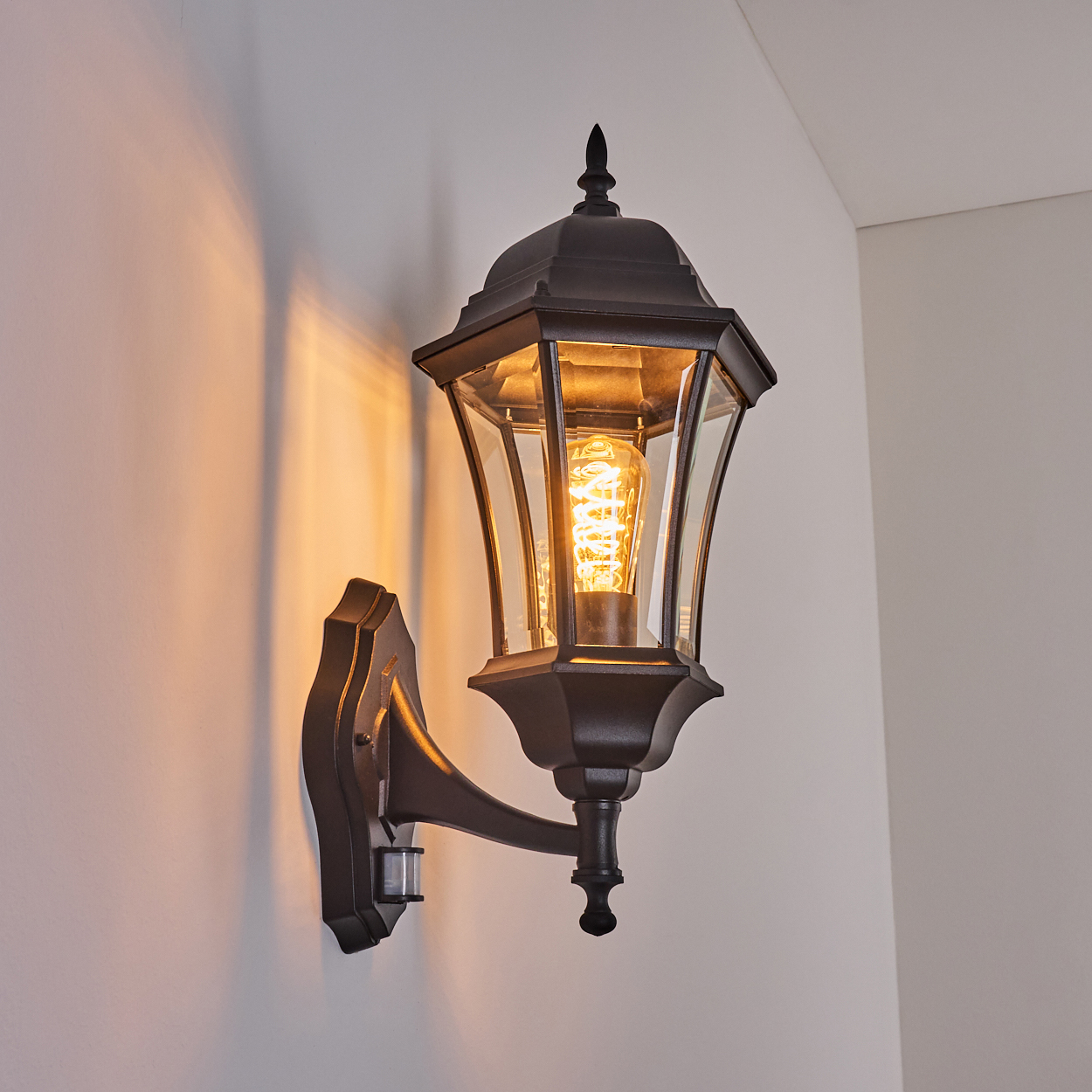 Schwarze Wandlaterne mit leuchtender Glühbirne, die einen warmen Schein auf die weiße Wand wirft. Die Laterne hat Glasscheiben.
