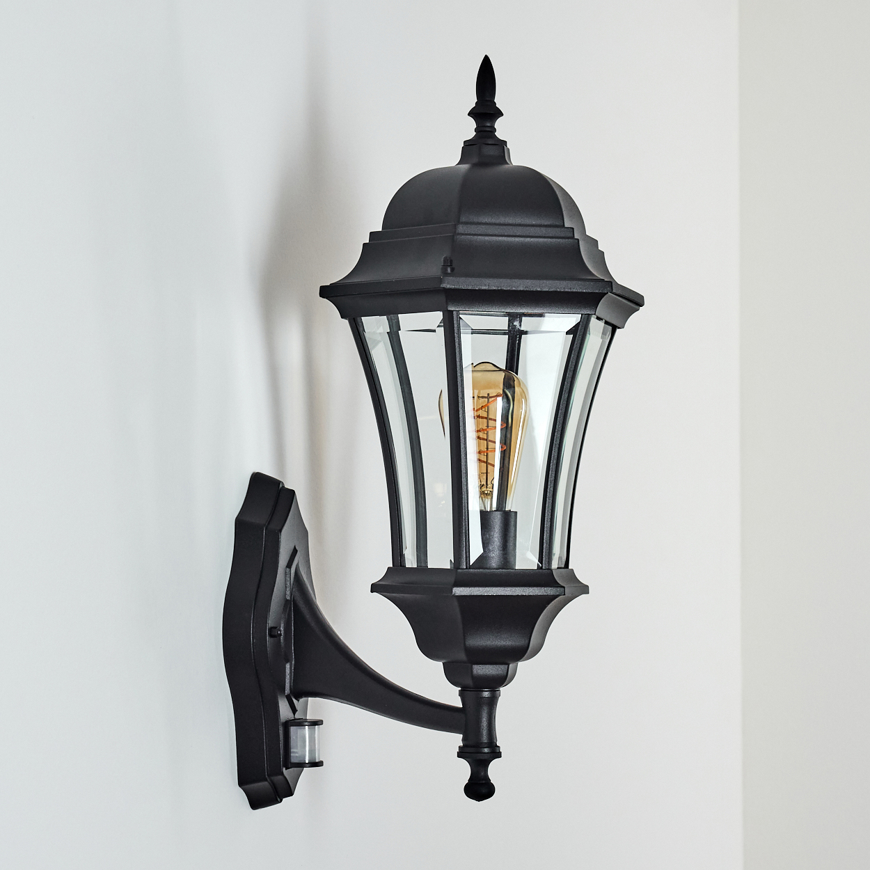 Schwarze Außenwandleuchte mit Glaspaneelen, an einer weißen Wand befestigt. Im Inneren der Lampe ist eine sichtbare Glühbirne. Ein Schatten fällt auf die Wand.