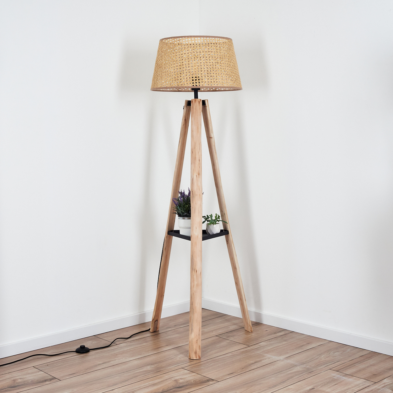 Eine Holz-Stativ-Stehlampe mit gewebtem Lampenschirm. Ein Regal hält Topfpflanzen. Der Boden ist aus Holz und die Wände sind weiß.