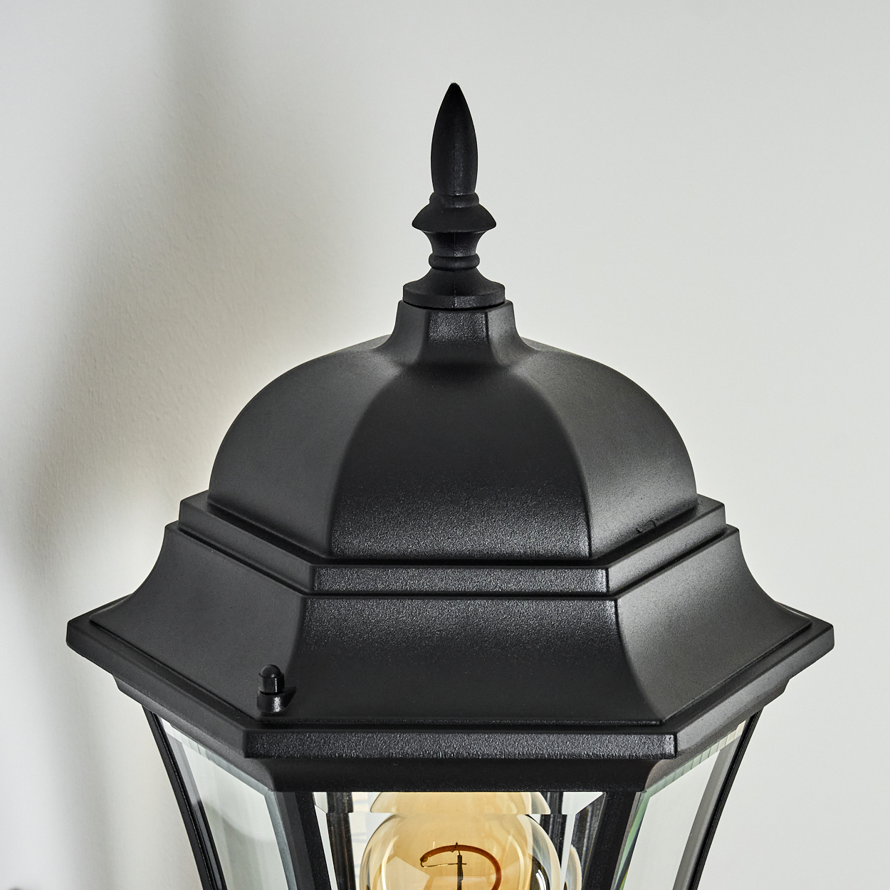 Nahaufnahme einer schwarzen Außenlampe. Sie hat eine spitze Spitze, ein kuppelförmiges Dach und Glasscheiben mit einer warmen Glühbirne.