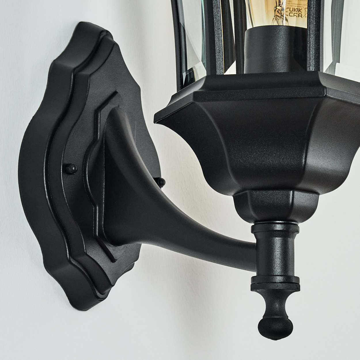 Eine schwarze Außenwandleuchte ist an einer weißen Wand montiert. Die Lampe hat ein Glasgehäuse und eine dekorative Halterung.